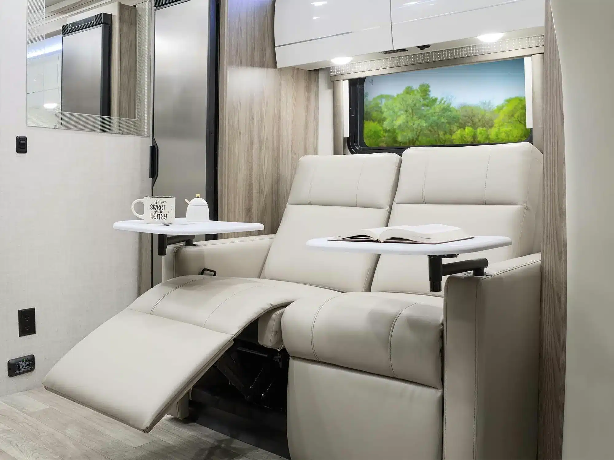 Innenraum des Thor Tiburon Sprinter mit beigem Relaxsofa, Klapptischen, Becher und Buch vor einem Fenster mit Ausblick ins Grüne.