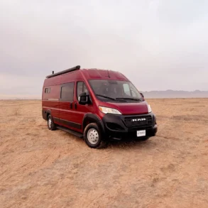 Burgunderroter RAM Promaster 3500 High Roof Kastenwagen von Trouvaille steht auf einem offenen, trockenen Feld mit Bergkulisse im Hintergrund
