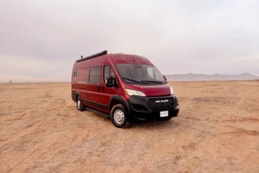 Burgunderroter RAM Promaster 3500 High Roof Kastenwagen von Trouvaille steht auf einem offenen, trockenen Feld mit Bergkulisse im Hintergrund