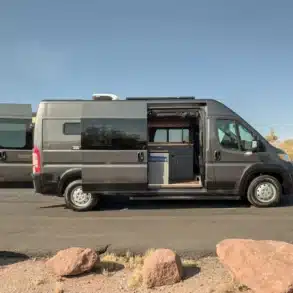 Seitenansicht eines ausgebauten RAM Promaster 2500 High Roof Kastenwagens mit geöffneten Türen, gezeigter Küche und Wohnbereich, stehend in wüstenartiger Landschaft.