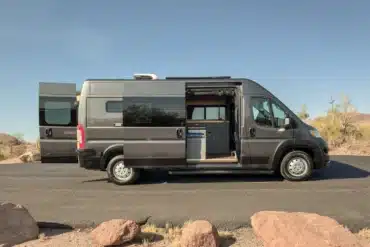 Seitenansicht eines ausgebauten RAM Promaster 2500 High Roof Kastenwagens mit geöffneten Türen, gezeigter Küche und Wohnbereich, stehend in wüstenartiger Landschaft.