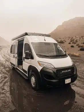 Weißer V’IBE RAM Promaster 2500 High Roof Campervan mit geöffneter Schiebetür in einer natürlichen Umgebung auf unbefestigtem Weg.
