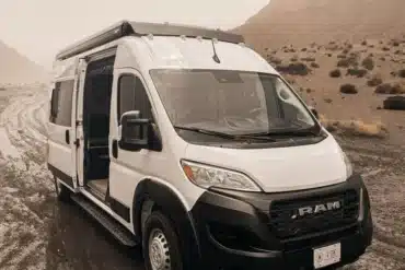 Weißer V’IBE RAM Promaster 2500 High Roof Campervan mit geöffneter Schiebetür in einer natürlichen Umgebung auf unbefestigtem Weg.