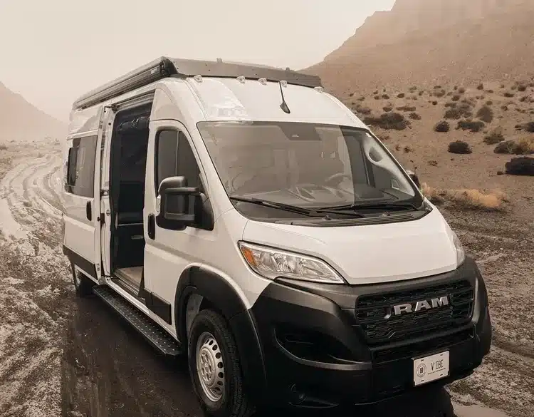 Weißer V’IBE RAM Promaster 2500 High Roof Campervan mit geöffneter Schiebetür in einer natürlichen Umgebung auf unbefestigtem Weg.