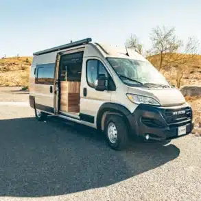Kompakter Vandiamo Kastenwagen-Camper auf RAM Promaster Basis mit geöffnetem Schiebetür, geparkt auf einem sonnigen Stellplatz in der Natur.
