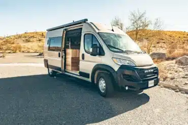 Kompakter Vandiamo Kastenwagen-Camper auf RAM Promaster Basis mit geöffnetem Schiebetür, geparkt auf einem sonnigen Stellplatz in der Natur.