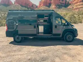 Whitney Camper Van auf Basis Ram Promaster 2500 High Roof, geöffnet am Standplatz vor roter Felskulisse, mit sichtbarer Küche und Innenausbau.