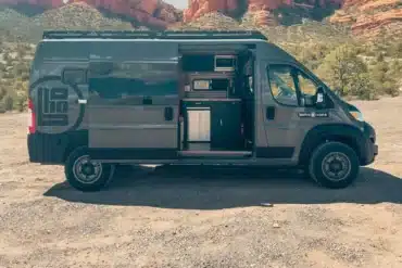 Whitney Camper Van auf Basis Ram Promaster 2500 High Roof, geöffnet am Standplatz vor roter Felskulisse, mit sichtbarer Küche und Innenausbau.
