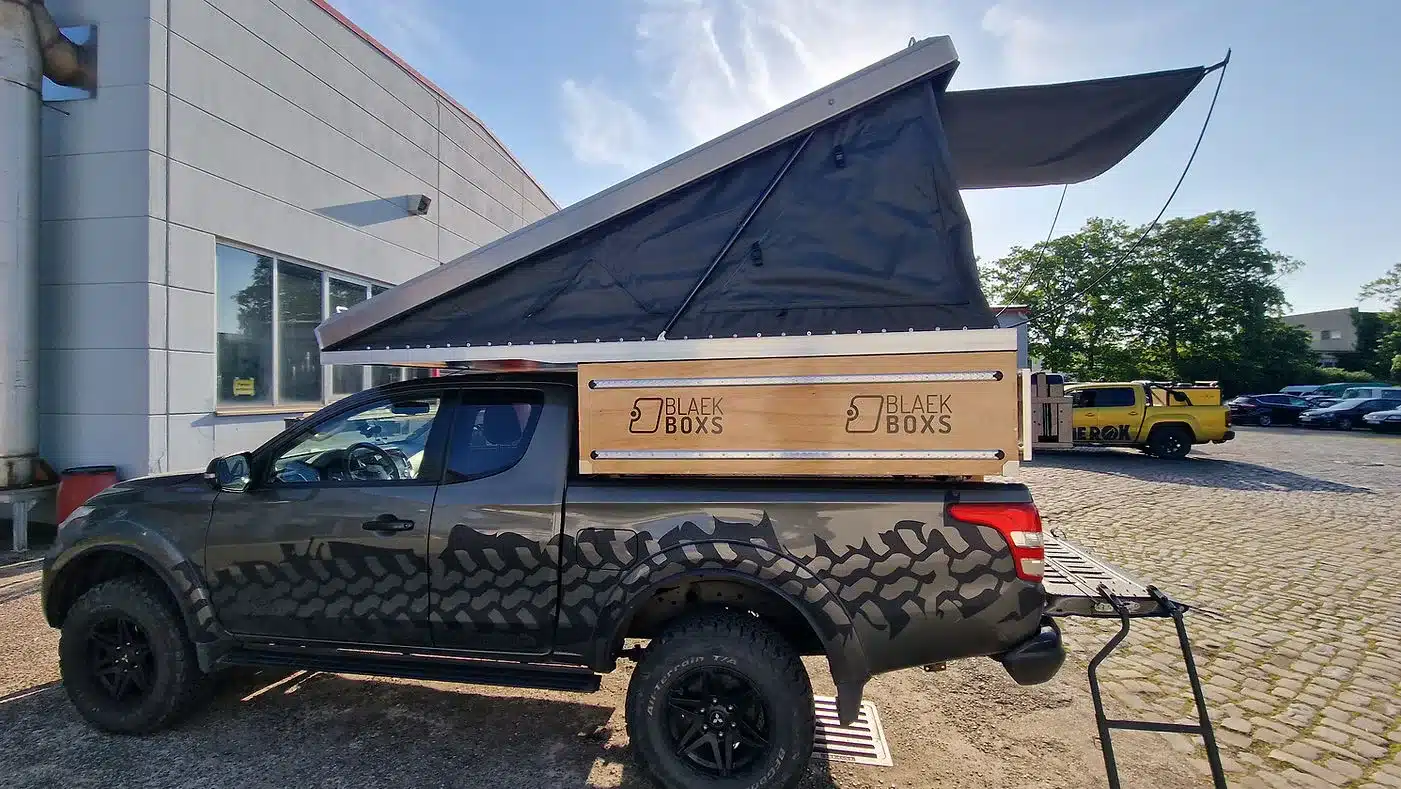 Pick-up mit montierter seitlicher Holzkiste und aufgeklapptem Dachzelt, Marke BLAEK BOXES, vor einem Gebäude