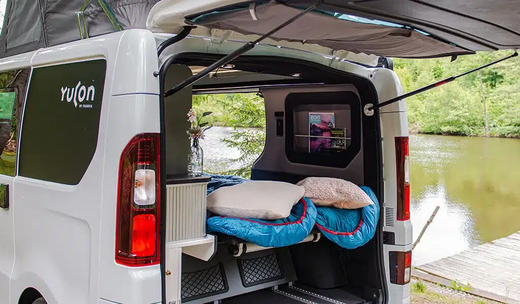 Yucon 55 SB FEVER Campervan mit geöffnetem Heck, aufgeklappter Schlafbank und Campingausstattung an einem Seeufer.