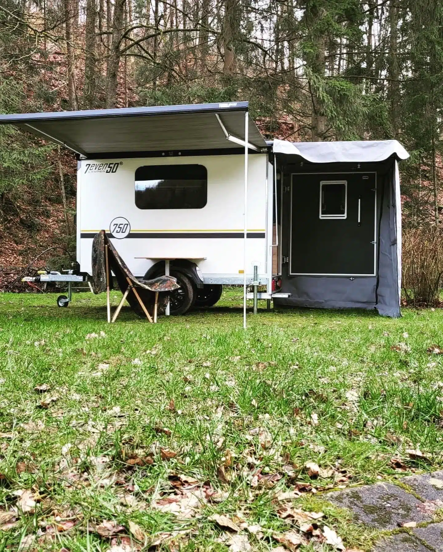 7even50 One Mini-Wohnwagen seitlich mit Markise und Campingstuhl auf einer Wiese vor Wald
