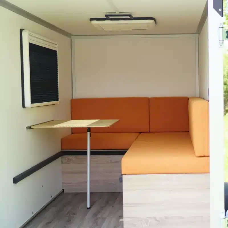 Innenraum eines Mini-Camper-Anhängers 7even50 Two mit orangefarbener Sitzgruppe und ausklappbarem Tisch