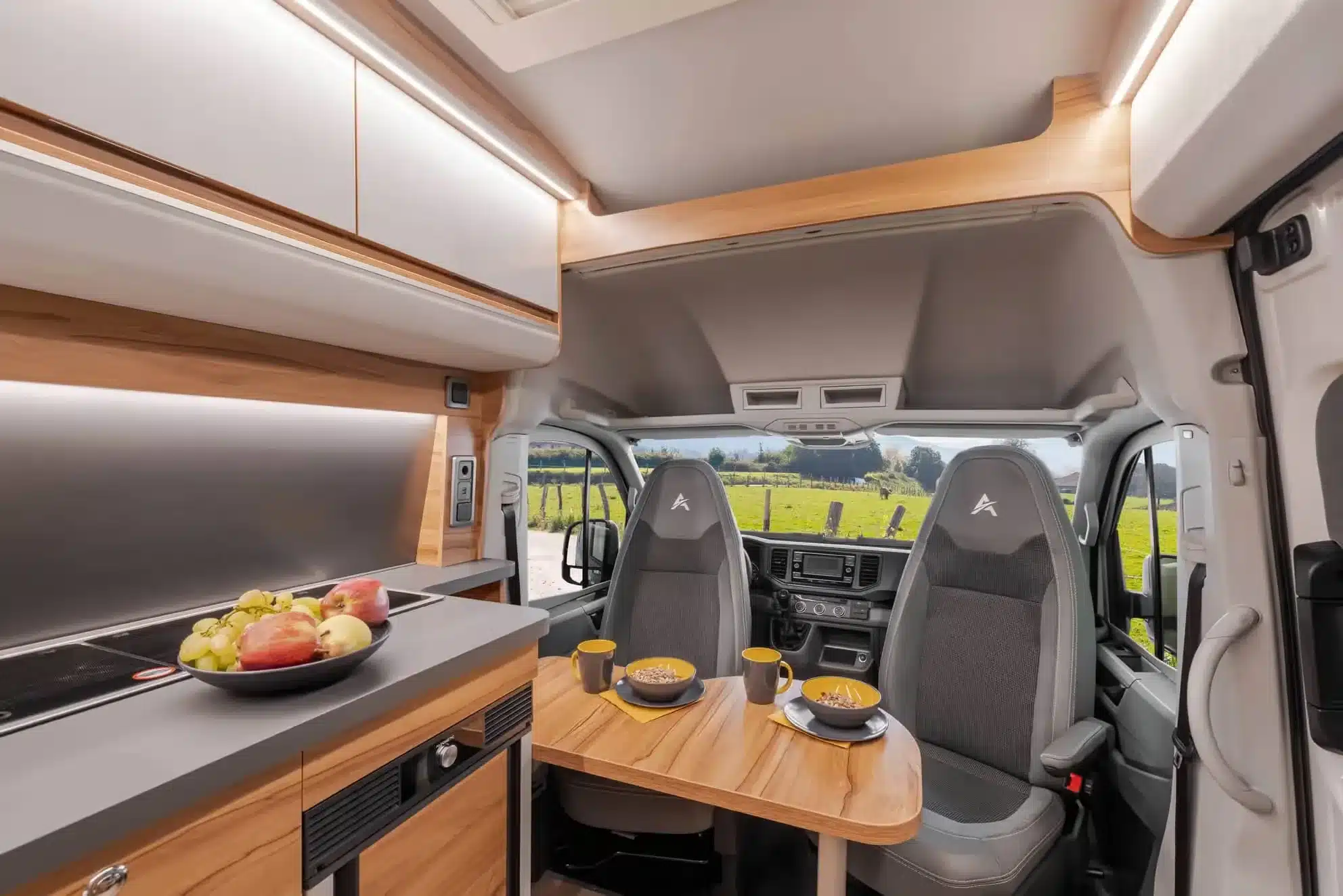 Wohnbereich und Küchenzeile im Affinity DUO M Campervan für zwei Personen, mit gedecktem Tisch und Ausblick ins Grüne.