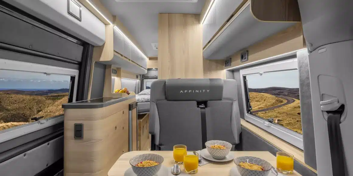 Innenraumaufnahme eines Affinity M Three Kastenwagens mit Sitzgruppe, Esstisch, moderner Küchenzeile und Bett im Heck.