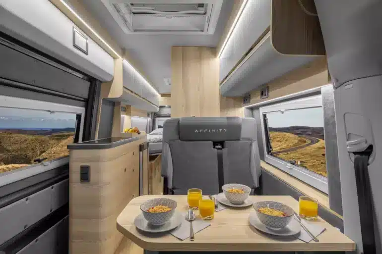 Innenraumaufnahme eines Affinity M Three Kastenwagens mit Sitzgruppe, Esstisch, moderner Küchenzeile und Bett im Heck.