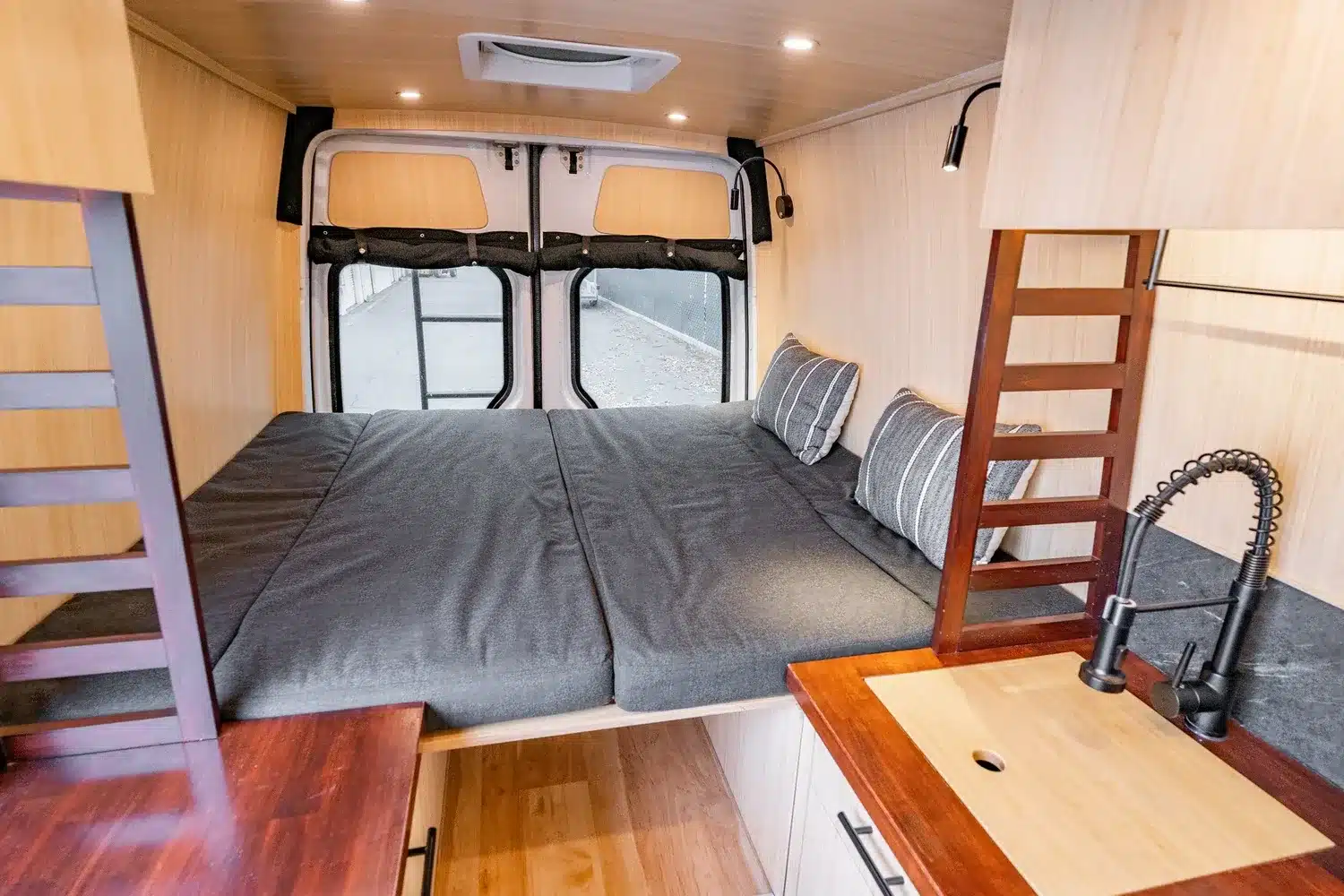 Innenraum von Appa Camper mit ausgezogenem Bett, Kissen und Küchenzeile aus Holz mit Wasserhahn.