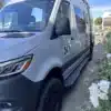 Baja Basecamp Campervan, silberner 4x4-Sprinter mit Offroad-Reifen, Dachträger und LED-Zusatzscheinwerfern, fotografiert im Freien.