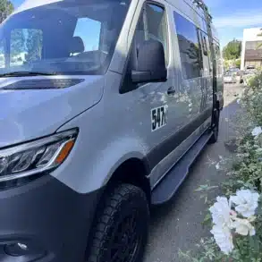 Baja Basecamp Campervan, silberner 4x4-Sprinter mit Offroad-Reifen, Dachträger und LED-Zusatzscheinwerfern, fotografiert im Freien.