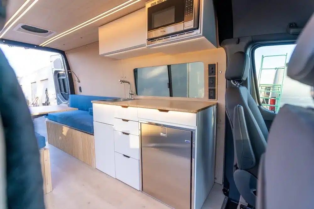 Innenansicht des Base Camp 2 Vans mit modularer Küche, Edelstahlkühlschrank, Mikrowelle, Spülbecken, Sitzbank und hellem Interior-Design