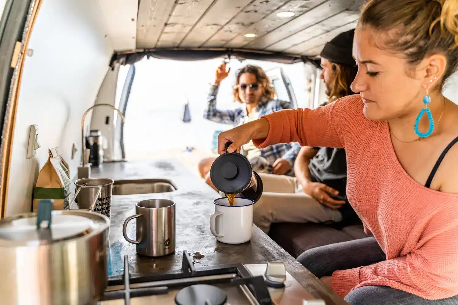 Frau bereitet Kaffee in der Camperküche von Blanca zu, im Hintergrund zwei weitere Personen im Van.