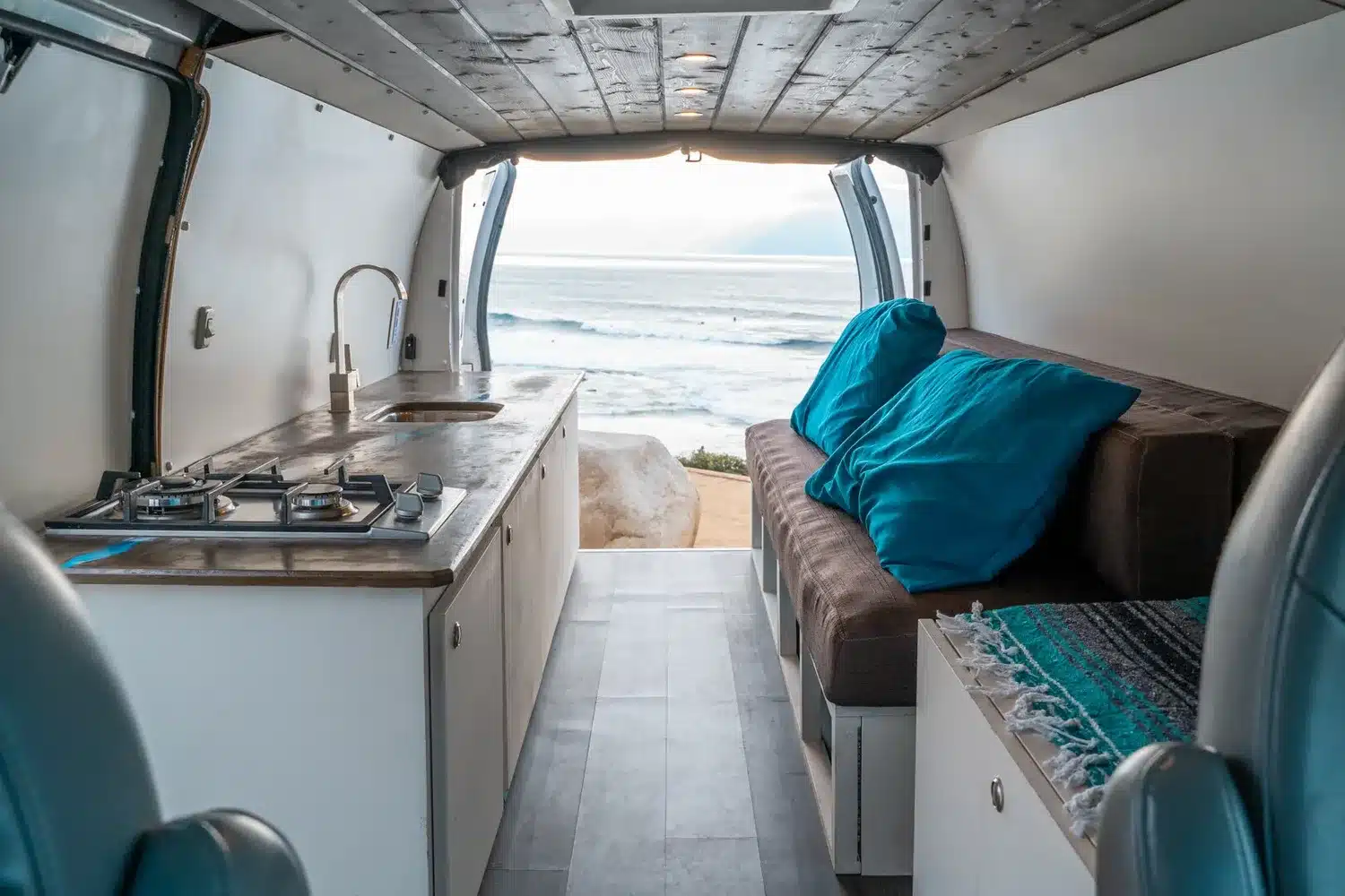Innenansicht eines ausgebauten Campervans mit Küchenzeile, Gasherd, Spüle und Sitzbank vor geöffneter Heckklappe mit Meerblick.