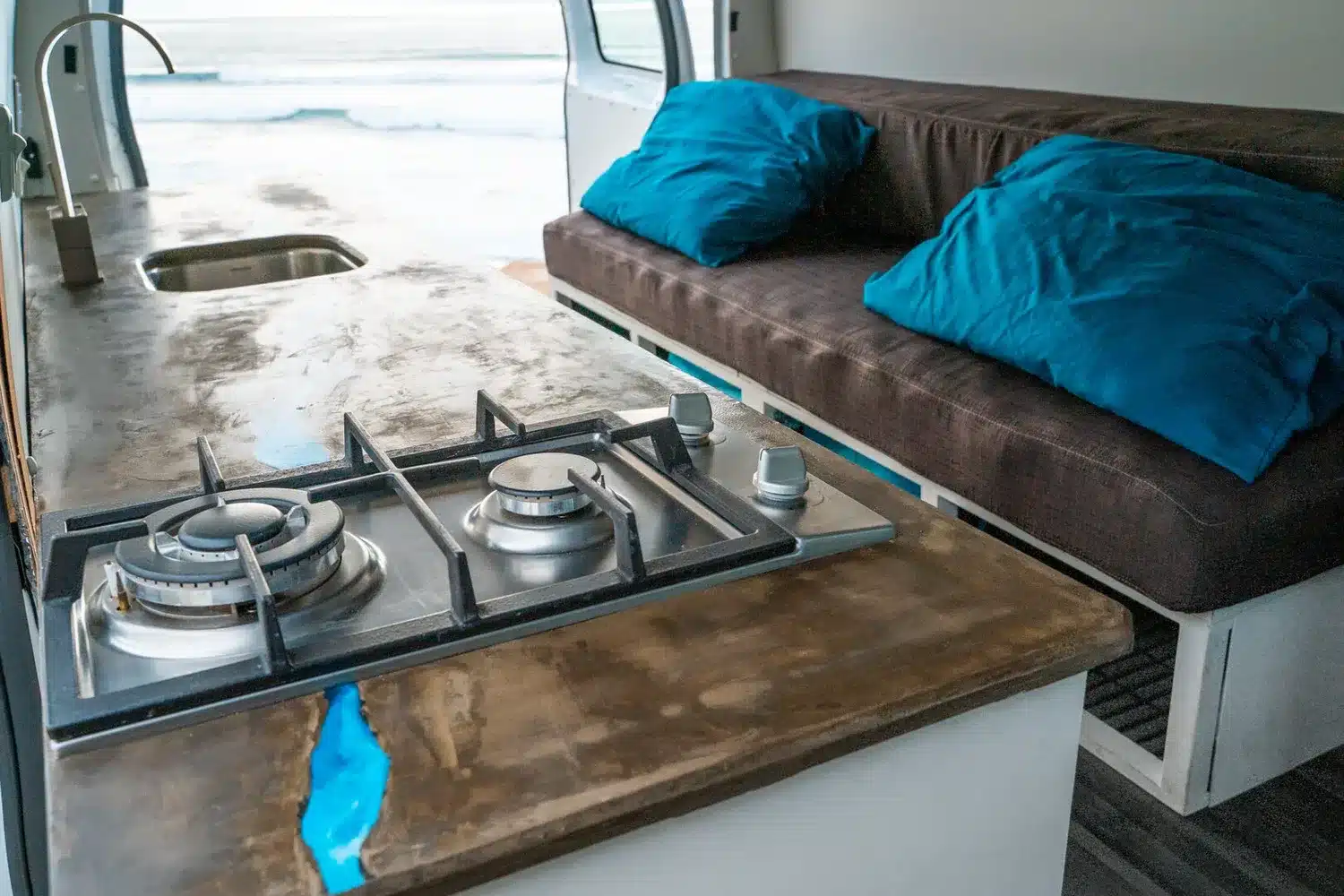 Camperküche mit Zweiflammen-Gaskocher, Beton- und Epoxy-Arbeitsplatte sowie Sitzbank mit blauen Kissen im Van Blanca