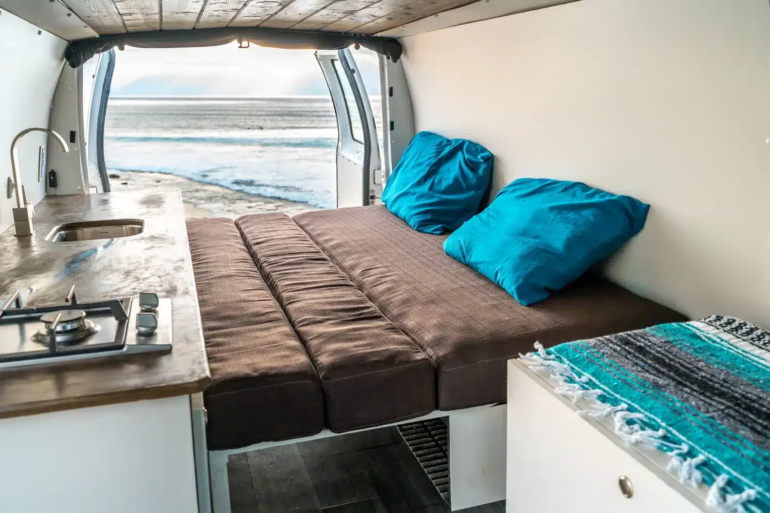 Ausgebauter Campervan Blanca mit ausgezogenem Bett und Küchenzeile, Türen geöffnet zum Meer.