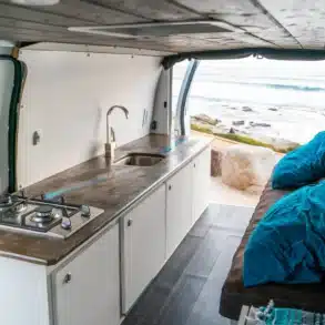 Campervan Blanca mit Arbeitsplatte in Betonoptik, eingebautem Gaskocher, Spüle und Sitzbank mit blauen Kissen, geöffnet mit Ausblick aufs Meer.