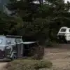 Zwei Camppass CR550 Offroad-Caravans stehen im Wald, eines wird von einem Geländewagen gezogen, das andere ist abgestellt, umgeben von Bäumen und Bergen.