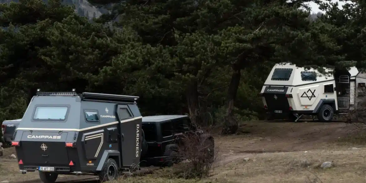 Zwei Camppass CR550 Offroad-Caravans stehen im Wald, eines wird von einem Geländewagen gezogen, das andere ist abgestellt, umgeben von Bäumen und Bergen.