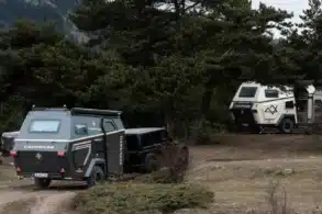 Zwei Camppass CR550 Offroad-Caravans stehen im Wald, eines wird von einem Geländewagen gezogen, das andere ist abgestellt, umgeben von Bäumen und Bergen.