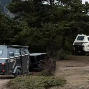 Zwei Camppass CR550 Offroad-Caravans stehen im Wald, eines wird von einem Geländewagen gezogen, das andere ist abgestellt, umgeben von Bäumen und Bergen.