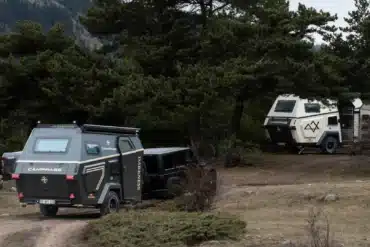 Zwei Camppass CR550 Offroad-Caravans stehen im Wald, eines wird von einem Geländewagen gezogen, das andere ist abgestellt, umgeben von Bäumen und Bergen.