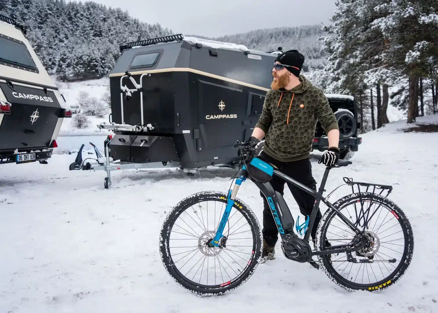 Ein Camppass CR550 Wohnanhänger steht im verschneiten Gelände, daneben ein Mann mit Fahrrad.