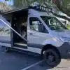 Carpool Cruiser Sprinter mit ausgefahrener Markise, Dachträger und Offroad-Stoßfänger auf einem Parkplatz.
