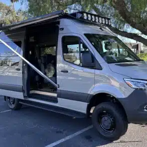 Carpool Cruiser Sprinter mit ausgefahrener Markise, Dachträger und Offroad-Stoßfänger auf einem Parkplatz.