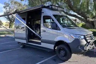 Carpool Cruiser Sprinter mit ausgefahrener Markise, Dachträger und Offroad-Stoßfänger auf einem Parkplatz.