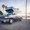Blauer Cobalt Cruiser Campervan von Vamonos Vans auf einem Parkplatz in Kalifornien, mit Palmen und Wasser im Hintergrund.