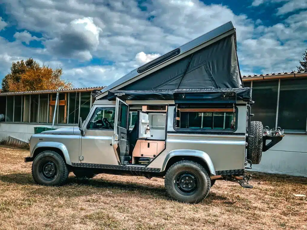 Land Rover Defender 110 Reisemobil mit Hubdach und Campingausbau auf einer Wiese vor einem Gebäude.