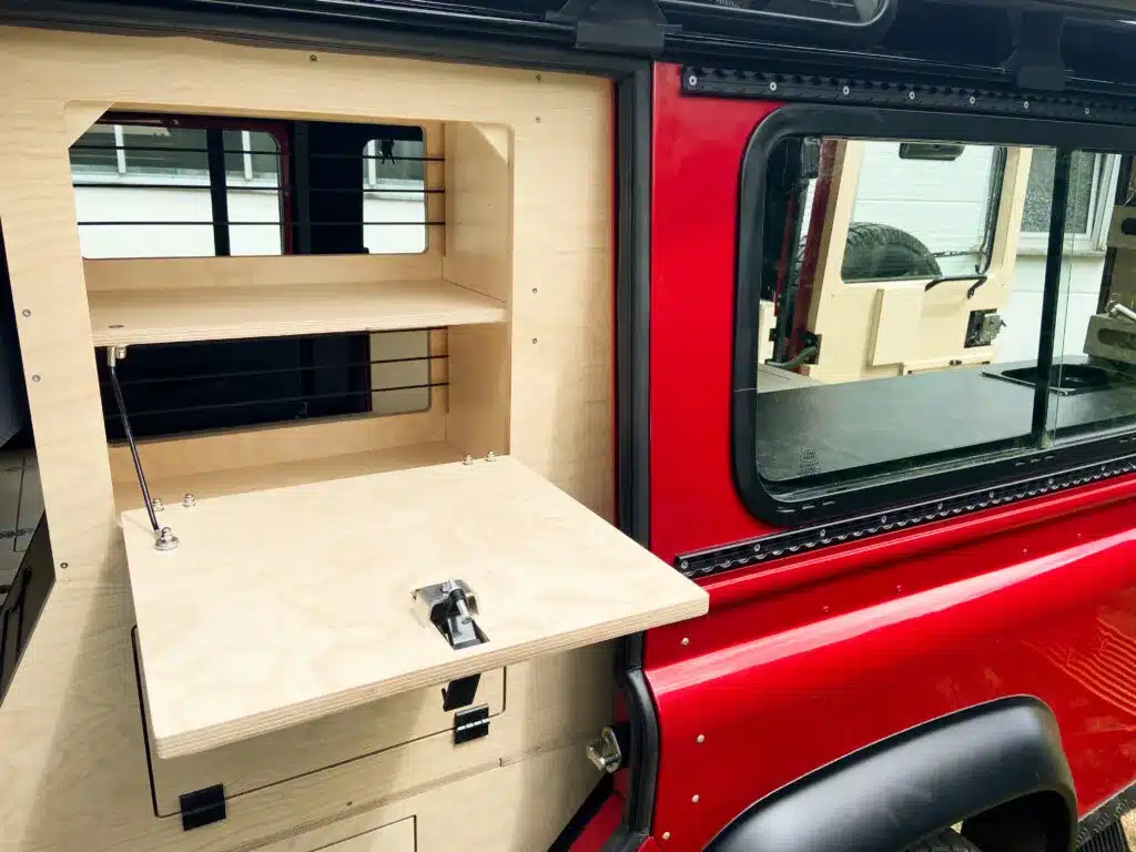 Geöffnetes Außenstauraumfach aus Holz am roten Defender 110 Alpenlandy mit Blick auf Innenausbau