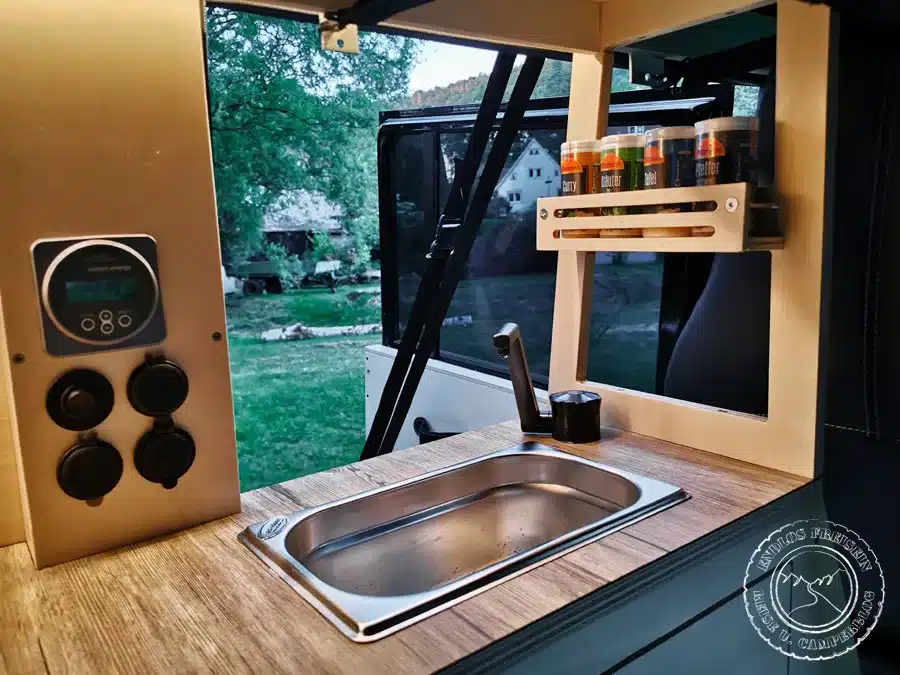 Küchenzeile mit Edelstahlspüle, Armatur, Gewürzregal und Bedienpanel im Camper-Ausbau eines schwarzen Land Rover Defender 110 SW.