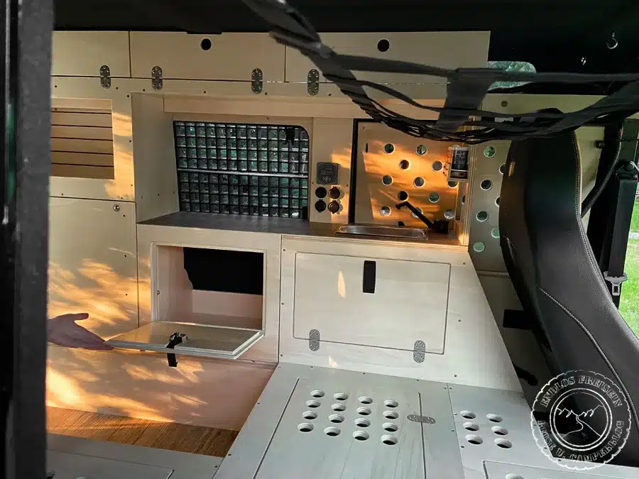 Innenraum eines zum Camper ausgebauten Land Rover Defender 110 SW mit heller Holzeinrichtung, Spülbecken, Klapptisch und Stauraum