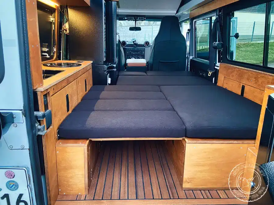 Innenausbau des Land Rover Defender 110 mit schwarzem Polster-Notbett, Küchenmodulen und Holzboden.