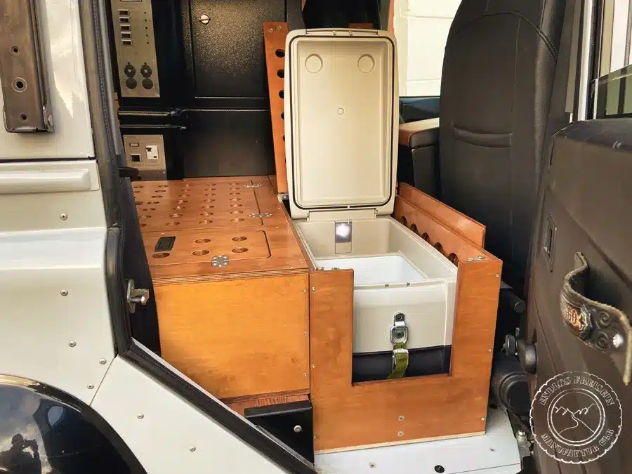 Geöffnete integrierte Kühlbox hinter dem Beifahrersitz im Land Rover Defender 110 Camper-Ausbau, umgeben von Holzmodulen.