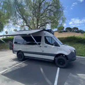 Adventure-Van „The Bear“ auf Mercedes Sprinter 144-Chassis mit ausgefahrener Markise auf Parkplatz