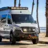 Mercedes Sprinter 144 4x4 Campervan mit Offroad-Umbauten und Zubehör, geparkt am Strand, mit Palmen im Hintergrund.