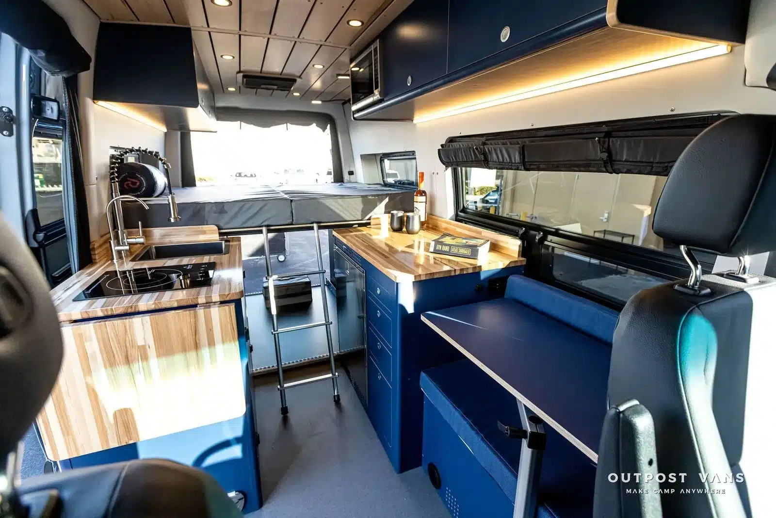 Küchenbereich, Sitzgruppe und Schlafplattform im Mercedes Sprinter 144 4x4 Camper Van ‚El Duque‘ mit Holzdekor und blauen Möbeln.