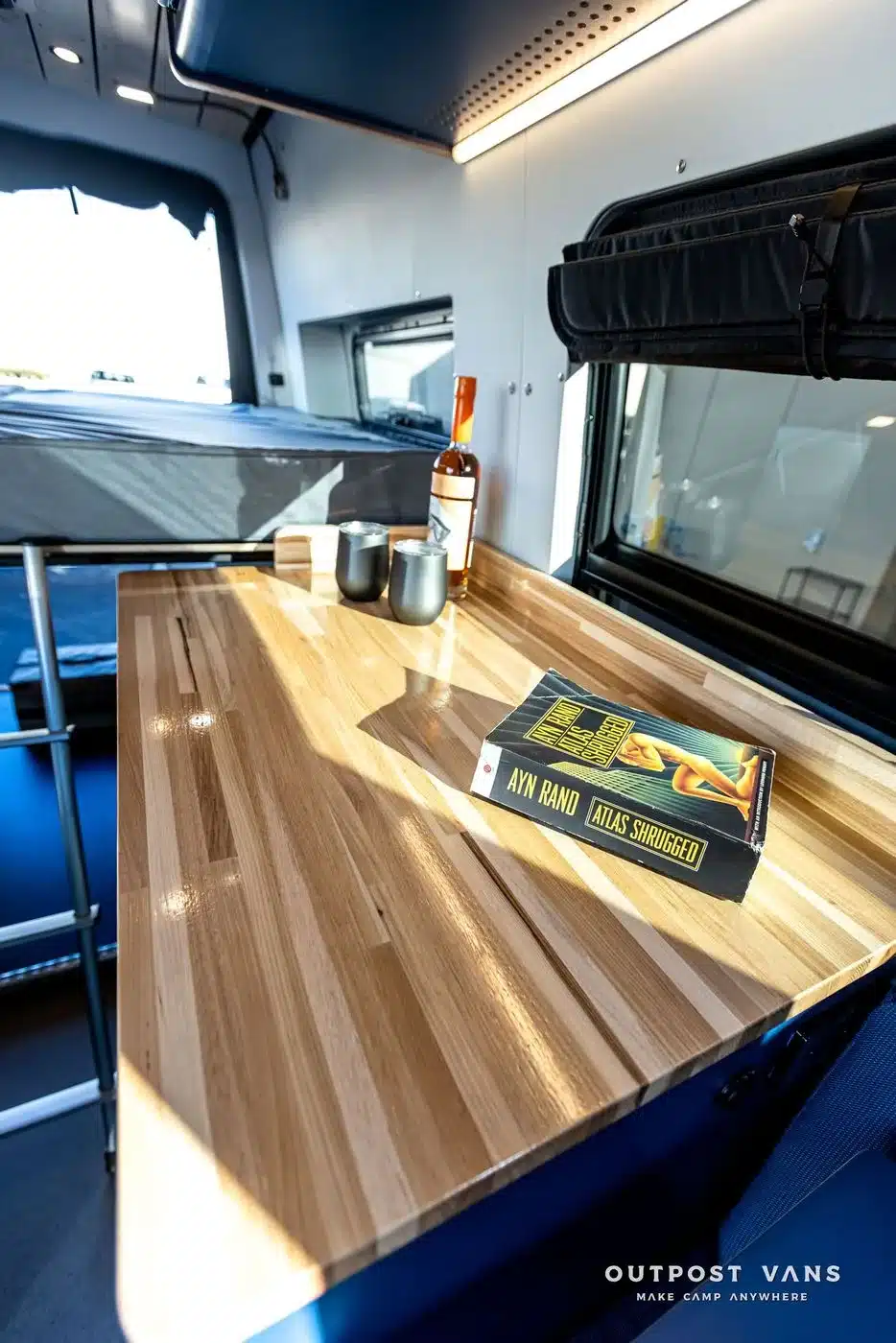 Küchenzeile mit heller Hartholz-Arbeitsplatte, Tassen, Flasche und Buch im Innenraum eines ausgebauten Mercedes Sprinter Campervans