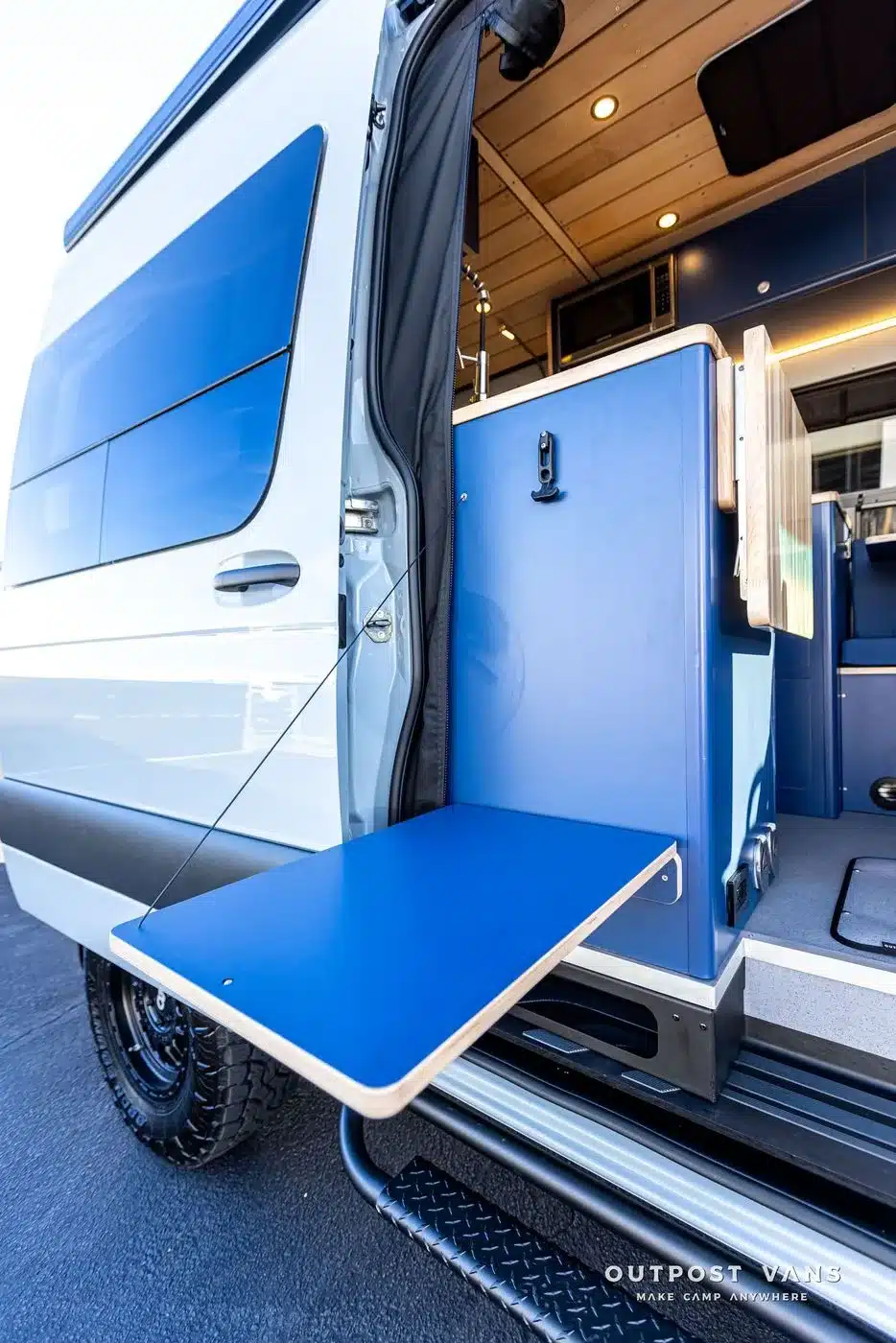 Ausklappbarer blauer Tisch an der Küchenzeile eines Mercedes Sprinter Campervans, Tür geöffnet, Innenausbau sichtbar.