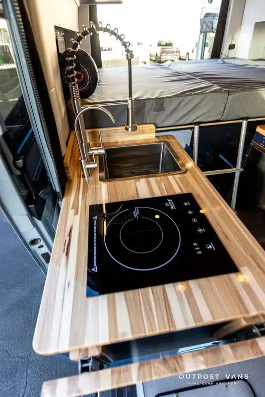 Küchenzeile im Mercedes Sprinter Camper mit Hartholz-Arbeitsplatte, Induktionskochfeld, Edelstahlspüle und flexiblem Wasserhahn.